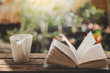 White cup of coffee and open book on the wooden table with the morning light and blurred nature in the garden background.