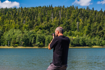 Fototapeta premium Adult man in hoodie walking around lake with DSLR camera ans shooting, beautiful summer day, half body portrait, landscape photography concept 