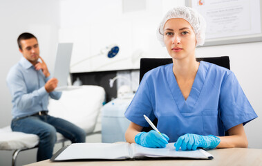 Portrait of young woman beautician sitting at desk writing notes while man patient checking result of procedure looking in mirror
