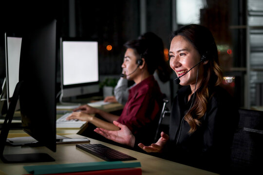 Happy smiling young beautiful Asian woman with headphones working at call center service desk consultant with her teammates at night, ready to take with customer on hands-free phone, happy workplace c