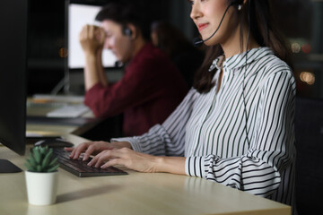 Hand typing on keyboard, Asian woman with headphones working at call center service desk consultant with her teammates at night, ready to chat or take with customer, happy workplace concept