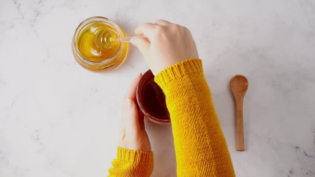 Close up of woman's hands in orange sweater adding honey into tea cup with immune boosting turmeric and ginger on white background. Top view. 4K
