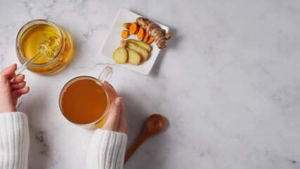 Close up of woman's hands in white sweater adding honey to tea cup with immune boosting turmeric and ginger on white background. Top view. 4K