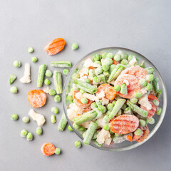 Frozen vegetables in glass bowl and on a table, green peas, carrot, cauliflower,  green beans, top view, square format