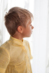 small boy looks into the distance through a brightly lit window behind a curtain