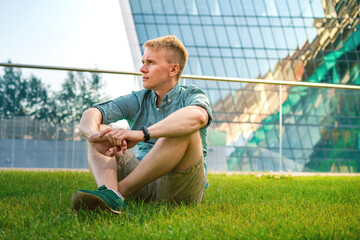 Naklejka premium A young man in a shirt rests on the grass in a business district with a view of glass skyscrapers