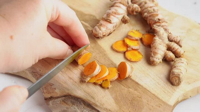 Close up of woman's hand cutting slices of turmeric root on wooden board. 4K