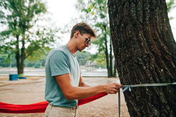 Portrait of a man in sunglasses and casual clothes hangs a hammock to a tree in the park with a concentrated face, knits slings.