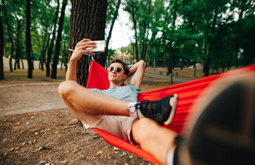 Young man in casual clothes taking a selfie while lying in the park on a hammock. Man resting on a hammock tied to a tree in the woods and uses a smartphone.