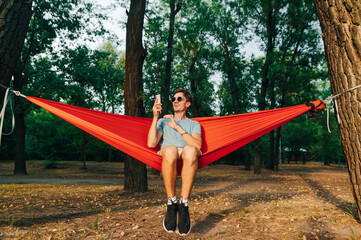 Happy stylish man in casual clothes swinging on hammock in forest with making video on smartphone with smile on face. A man swings on a hammock in the park and uses a smartphone.