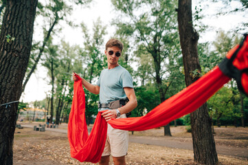 Handsome young man ties a red hammock to a tree in the park for relaxation.