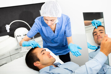 Confident woman cosmetologist examining male client face before procedure in esthetic clinic. Man looking at mirror.
