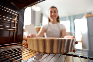 food cooking, culinary and people concept - young woman putting baking dish into oven at home...