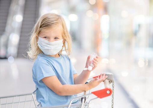 Baby Girl Wearing Medical Protective Facemask Applies Sanitizer For Cleaning Hands In Public Place During Coronavirus And Flu Outbreak Sits Inside Shopping Cart