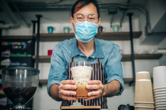 Business woner man wearing face mask for good hygiene coffee shop owner serving ice coffee young entrepreneur,friendly waitor with glasses smiling holding and serving.