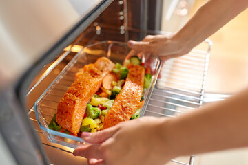 culinary, food and people concept - woman cooking salmon fish with vegetables in baking dish in oven at home kitchen