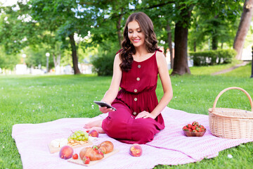 leisure and people concept - happy smiling woman with picnic basket and smartphone photographing food on blanket at summer park