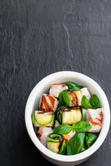 Grilled chicken and zucchini roll in clay bowl on black stone background