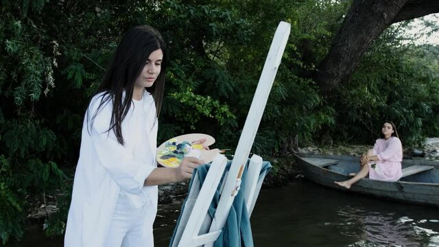 A painter paints a real landscape on the textile, a girl sitting in a boat on the shore of a lake. Young brunette woman draw a jacket.