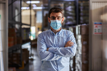 Fototapeta na wymiar Young successful attractive businessman with surgical mask on standing in warehouse with arms crossed and looking at camera. Corona outbreak concept.