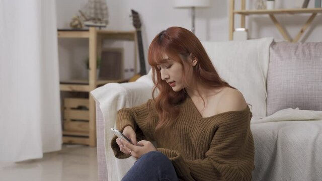 asian pretty woman is taking a pause to think while typing plans on her smartphone, drinking coffee in contemplation in the living room at home.