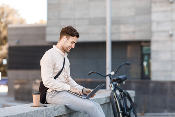 Freelancer working outside. Smiling guy in headphones with coffee surfing in laptop