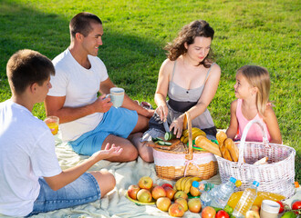 Happy young woman with her husband and two children enjoying picnic on green meadow together