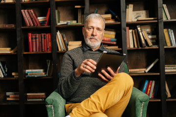 Portrait of bearded mature adult male browsing internet on digital tablet, sitting at home on background of bookshelves in cozy dark room with an authentic aristocratic interior, lookig at the camera.