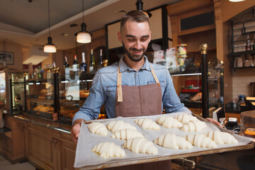 Male baker carrying tray of raw croissants to the oven at his bakery, copy space