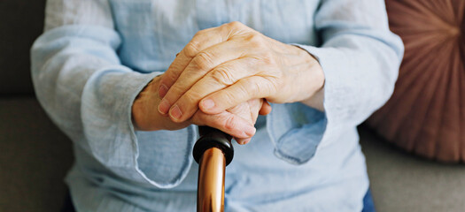 Elderly woman sitting in nursing home room holding walking quad cane with wrinkled hand. Old lady wearing beige cardigan, metal aid stick handle bar close up. Interior background, copy space
