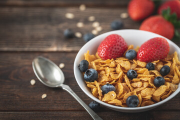 Corn flakes with blueberries and raspberries in white bowl in dark wooden desk.