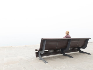 Woman in despair sitting on a lonely bench and looking at grey distant horizon on a ocean foggy day.