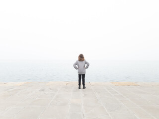 Girl child looking at grey distant horizon on a ocean foggy day.