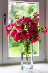 Rosenstrauß mit roten Rosen in Glasvase, auf Fensterbrett mit Blick hinaus in einen weiten Garten