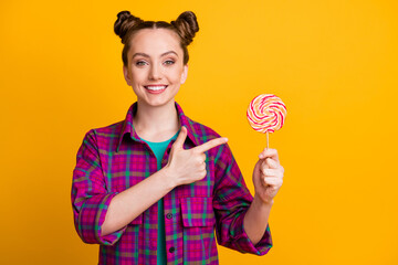 Portrait of her she nice attractive cheerful girl wearing checked shirt holding in hand demonstrating striped lollypop candy isolated bright vivid shine vibrant yellow color background