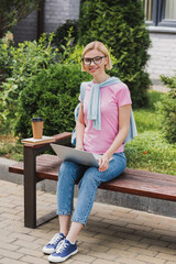 Fototapeta premium blonde student in glasses using laptop near book and paper cup while sitting on bench