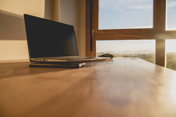 Laptop and mobile phone on a wooden table in a room with a window during dawn