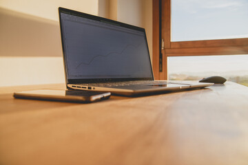 Laptop and mobile phone on a wooden table in a room with a window during dawn