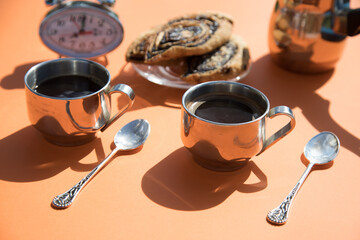 On an orange background on a Sunny morning, there are metal coffee cups, a coffee pot, buns on a plate, and an alarm clock. Horizontal orientation