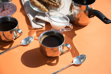 Coffee in metal cups, milk, rolls, orange background, sun glare and shadows-summer Breakfast....