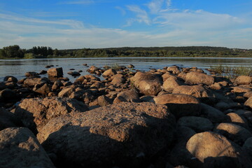 Rocky riverbank with large boulders in warm evening light. Calm water and forested hills in the background create a peaceful natural landscape