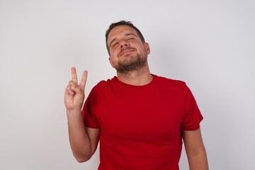 Young caucasian man wearing red t-shirt over white background smiling with happy face winking at the camera doing victory sign. Number two.