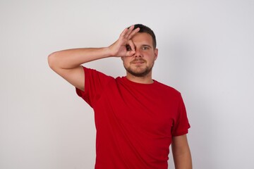 Young caucasian man wearing red shirt standing over isolated yellow background doing ok gesture with hand smiling, eye looking through fingers with happy face.