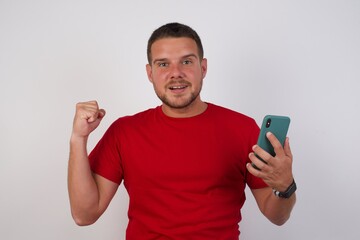 Positive  Young handsome Cucasian man wearing red shirt standing against white background holds modern cell phone connected to headphones, clenches fist from good emotions, exclaims with joy,