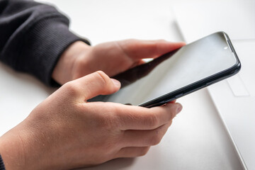European child hands holding black smartphone with fingers and black screen on white desk infront of white notebook at school for research and mobile gaming or game addiction of kids and grown ups