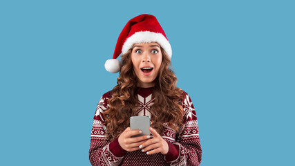 Millennial woman with cellphone shocked over huge Christmas discount in online shop, blue background