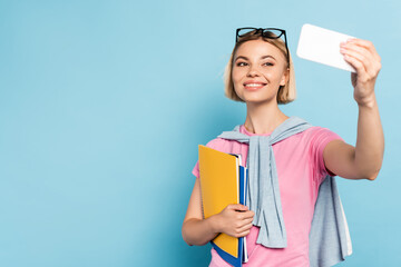 blonde student holding notebooks and taking selfie on blue