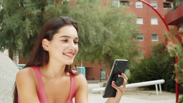 Happy young latin woman talking and texting on smart phone, wearing casual clothes sitting on bench. Generation z students life, girl online shopping near campus, social media and video conference.