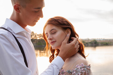 A beautiful wedding couple on the riverbank at sunset. A woman in a gray dress with flowers, a man in a suit and suspenders hug each other, love. Lovers on a walk