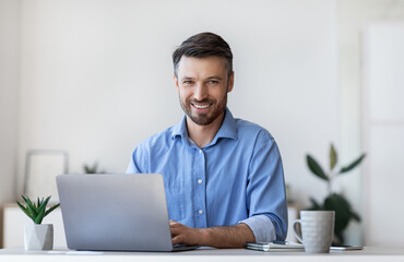 Smiling Young Businessman Sitting At Desk With Laptop In Modern Office